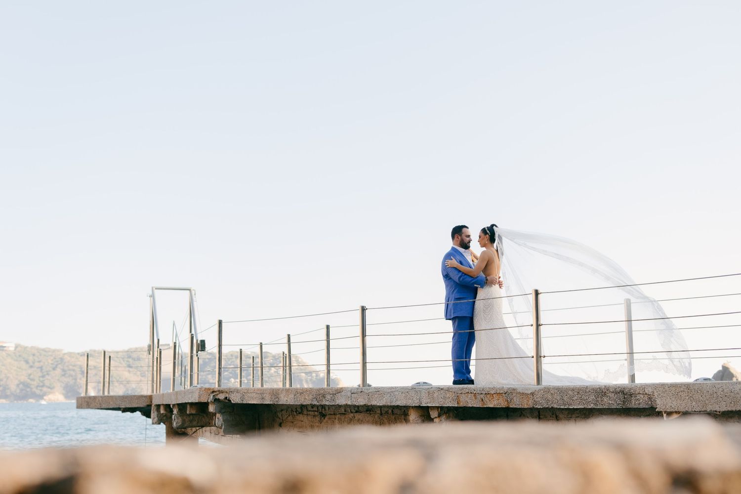 Newlyweds share intimate moment on wooden pier overlooking waterfront.