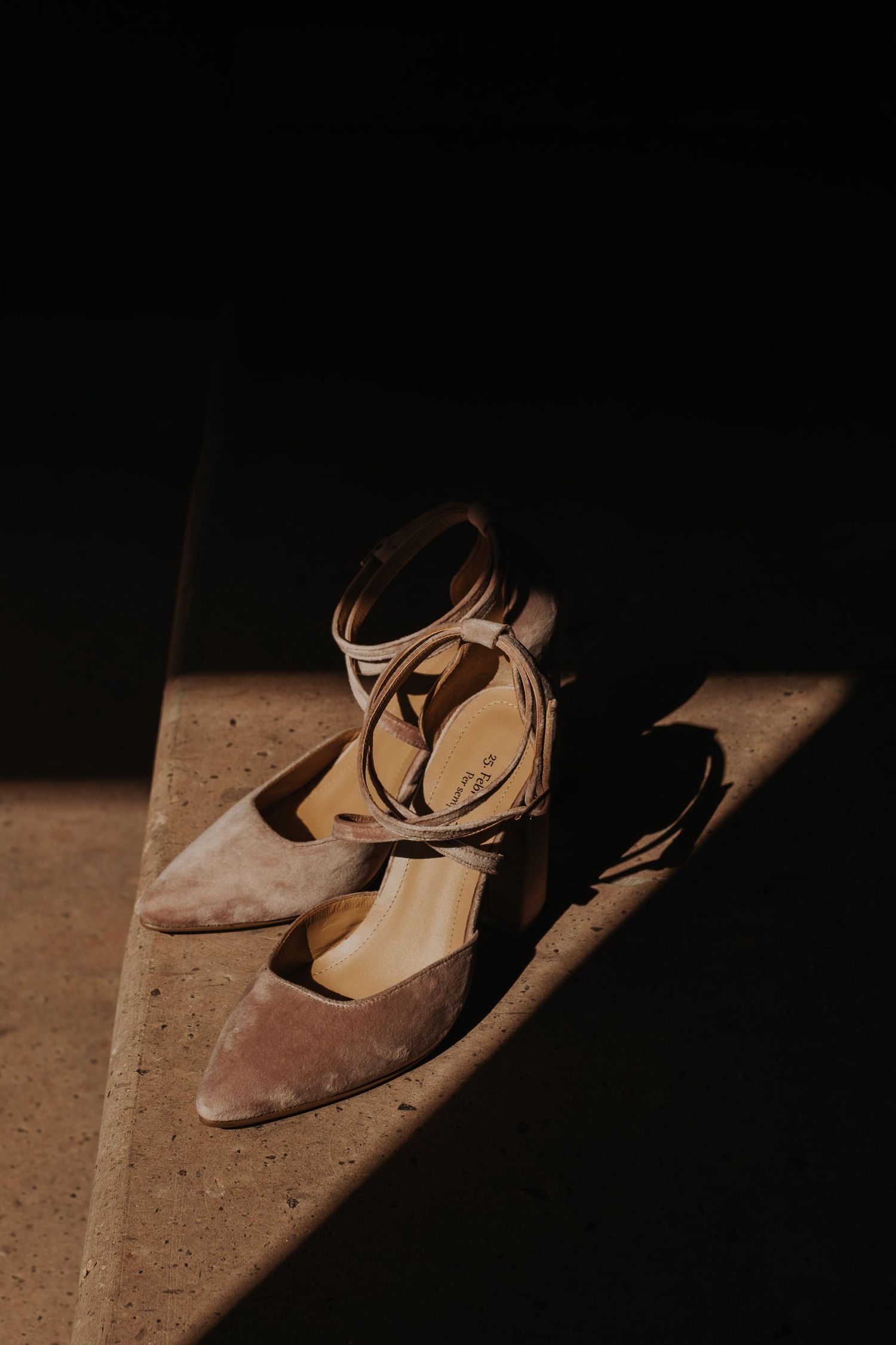 Elegant beige ankle-strap heels illuminated by dramatic sunlight on a textured surface.