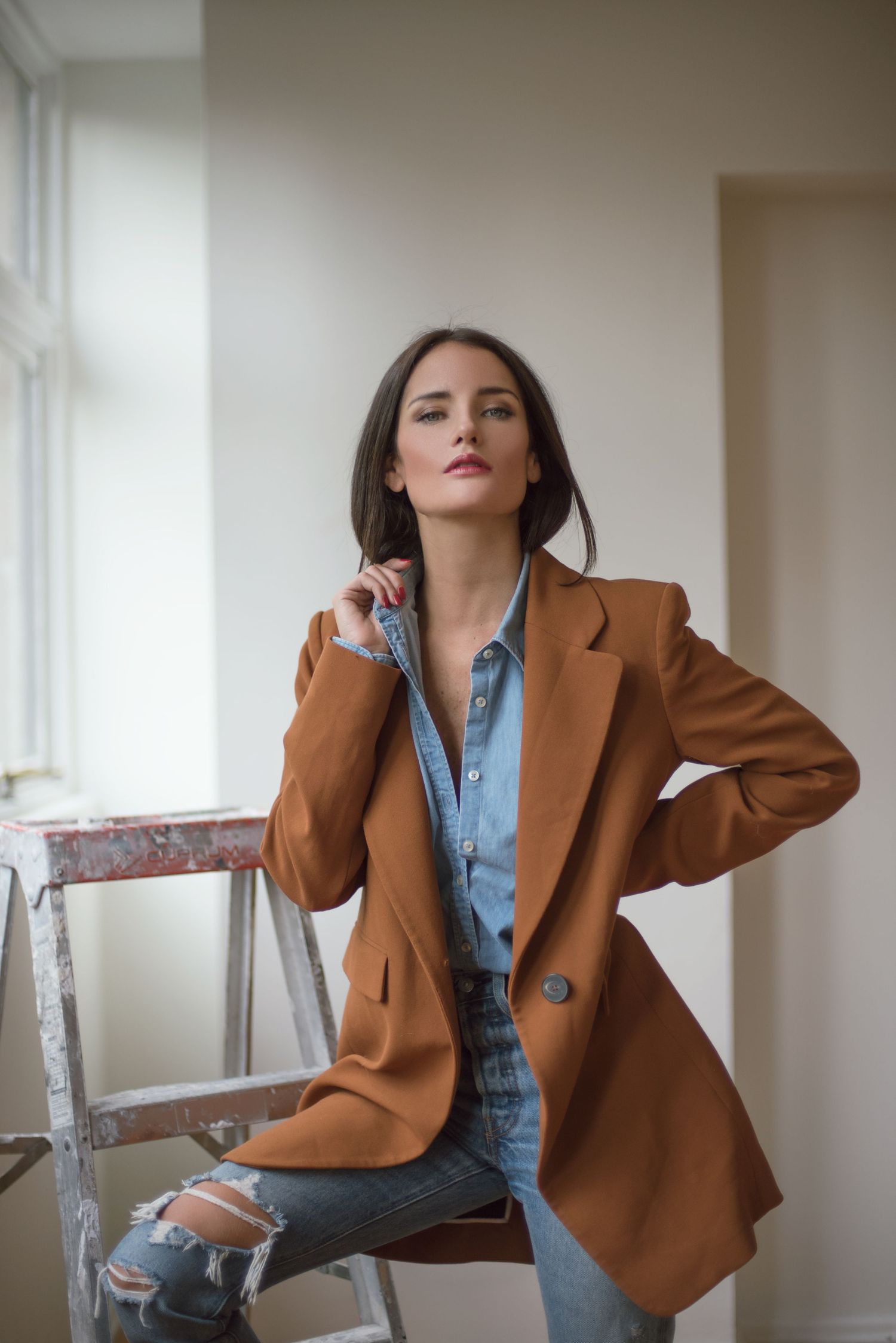 Fashion model in brown blazer and ripped jeans poses against white wall near window.