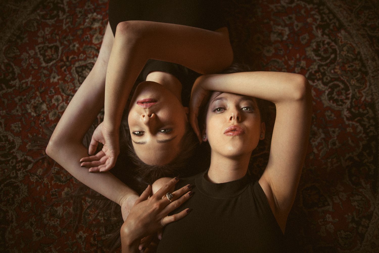 Artistic overhead portrait of two people lying on patterned carpet in dark room.