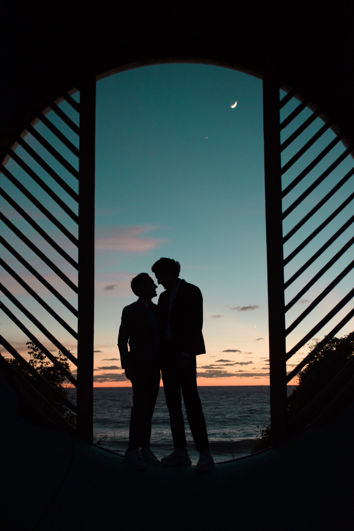 Silhouetted couple standing in geometric doorway at sunset with crescent moon and ocean view.