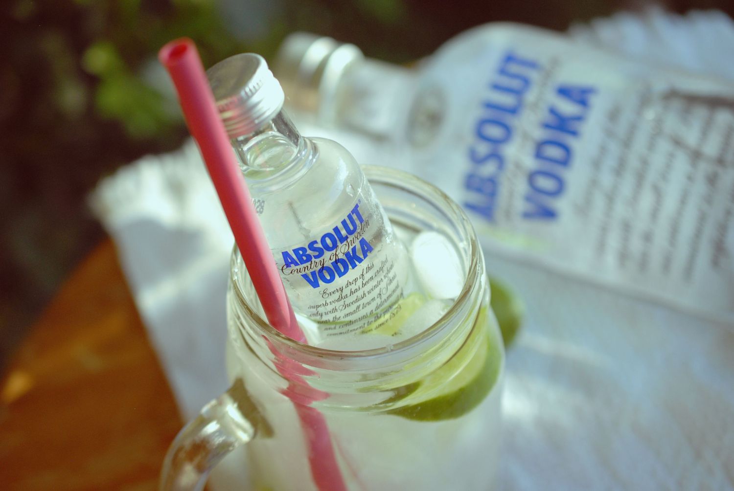 A bottle of Absolut Vodka with a pink straw against a blurred background with natural lighting.