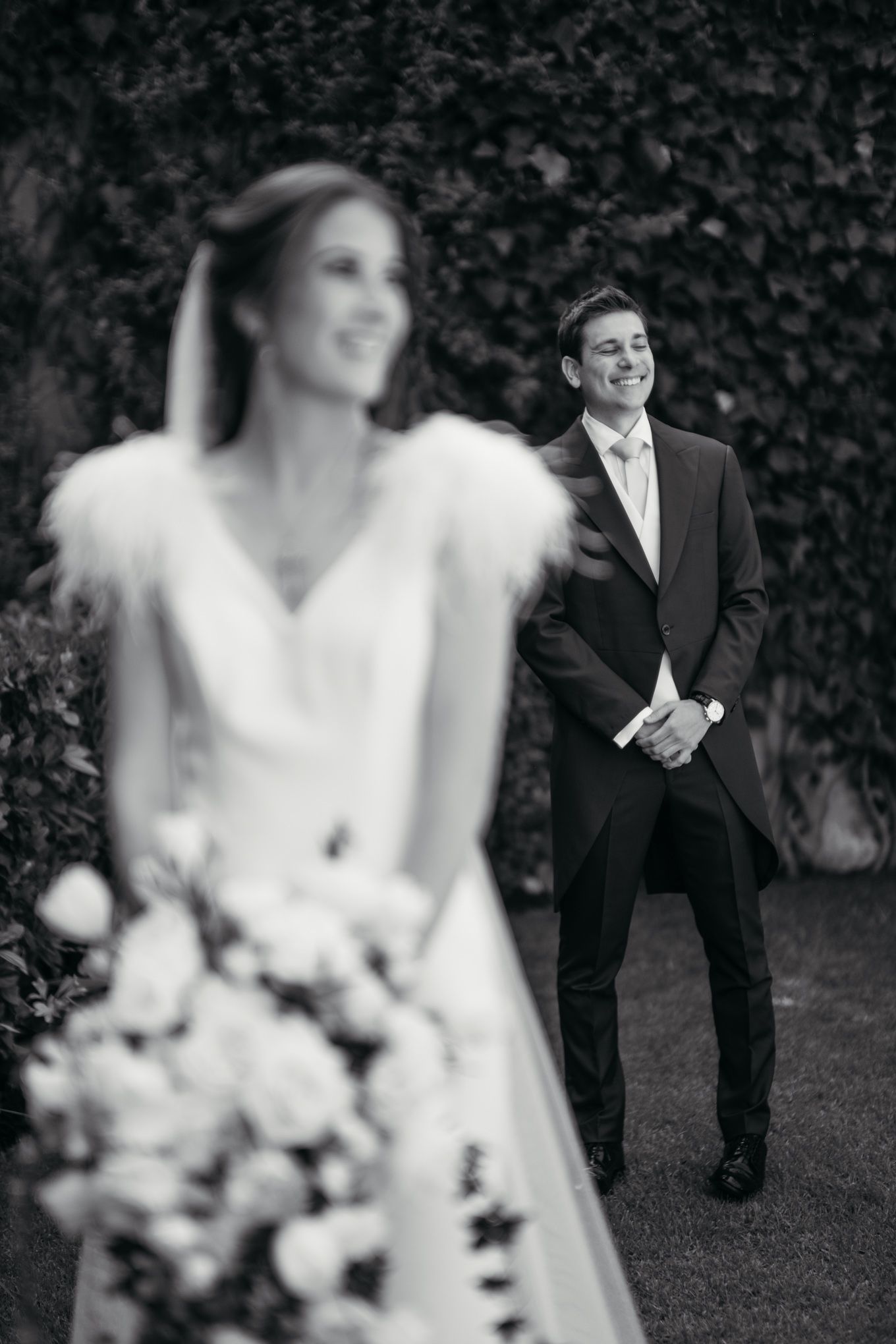 Black and white wedding photo with bride holding bouquet in foreground while groom stands in background by ivy wall.
