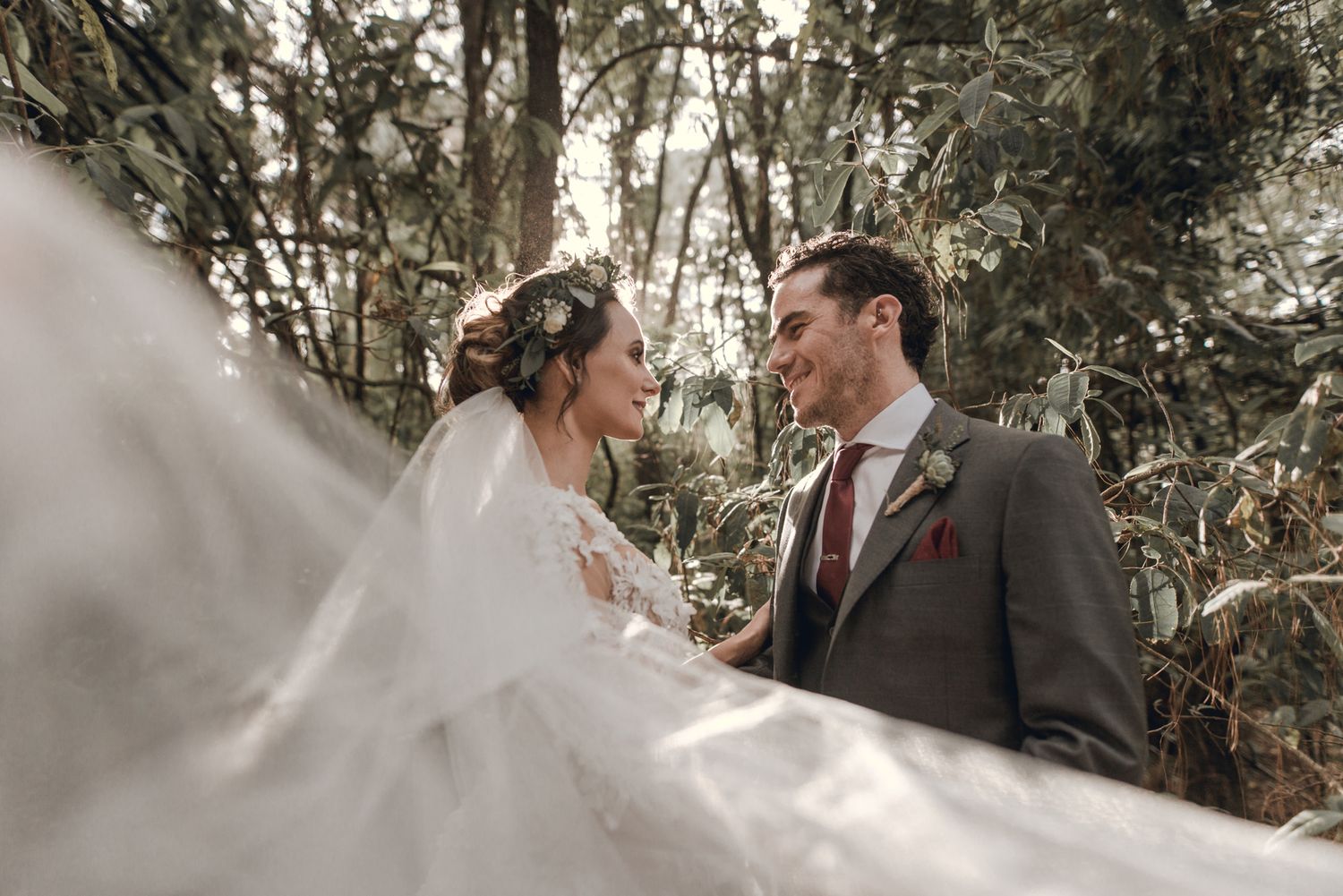 A bride in flowing white dress and veil gazes lovingly at her groom in brown suit among trees during outdoor wedding.