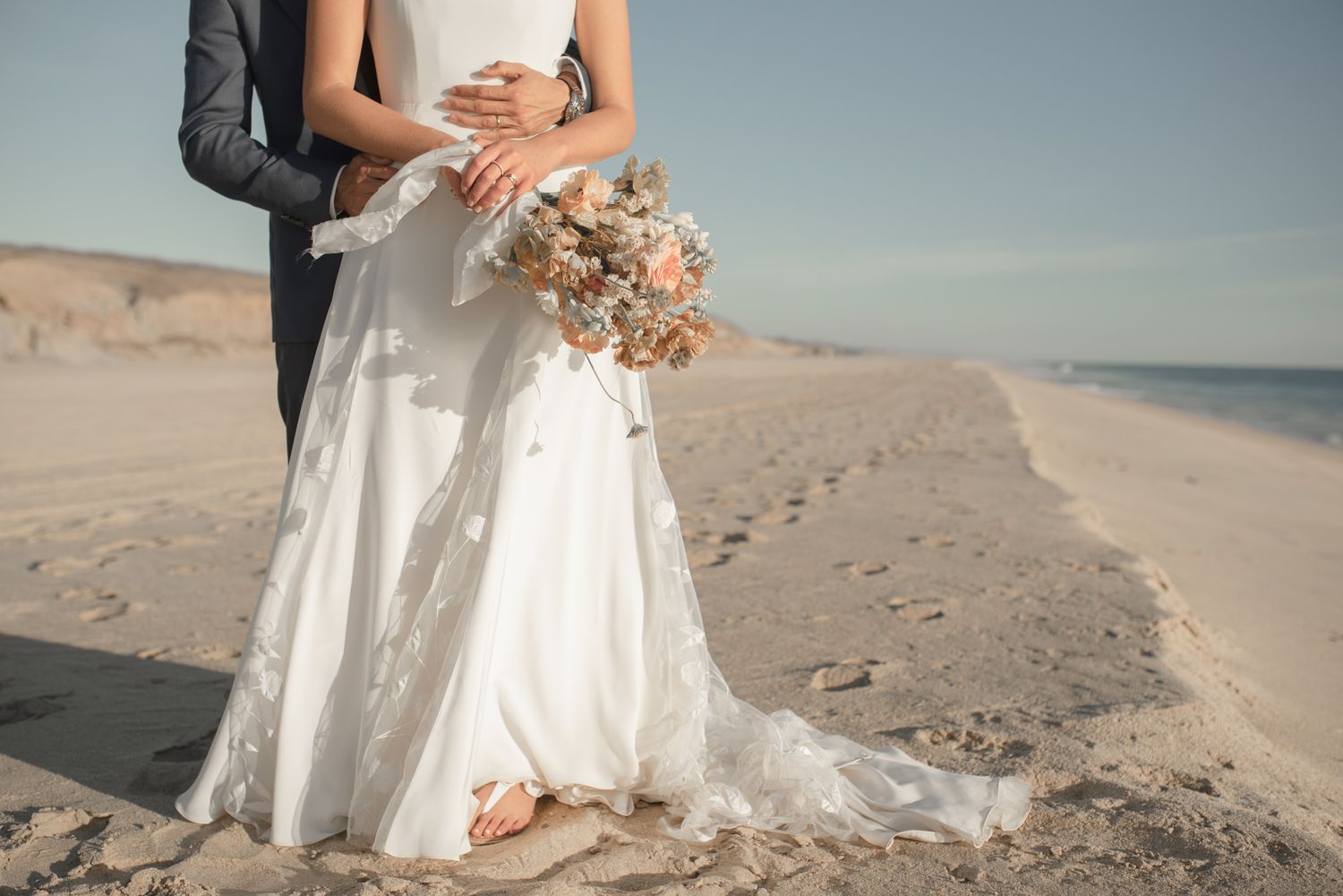 A couple embraces on a sandy beach, with the bride in a flowing white dress holding a pastel bouquet at sunset.