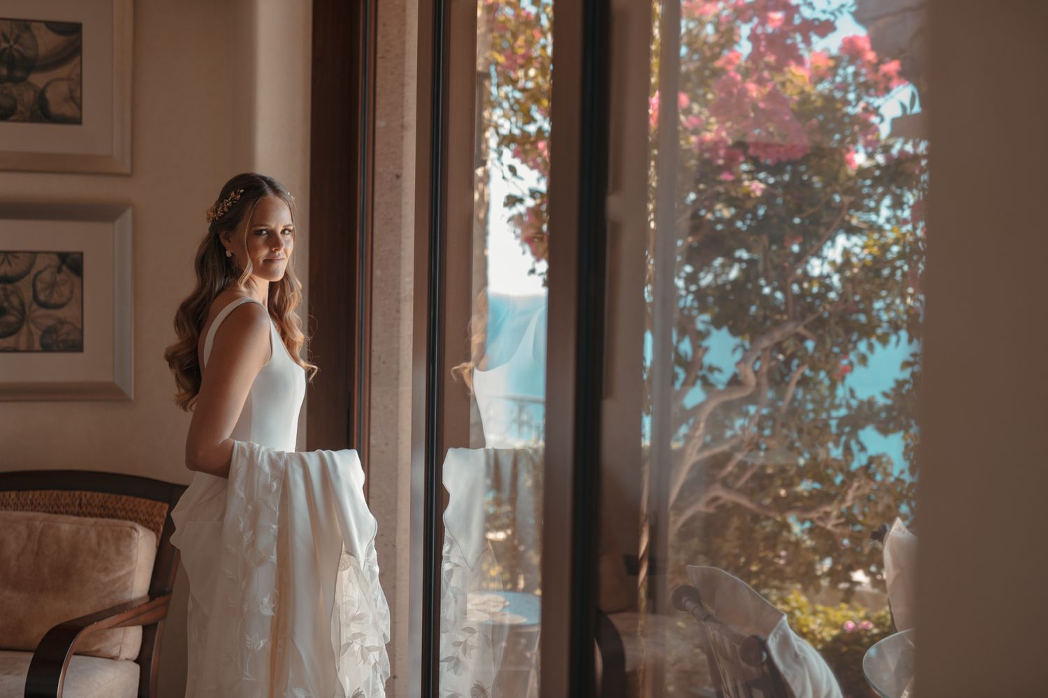 Soft natural light streams through window onto elegant white wedding dress while figure gazes at scenic outdoor view.