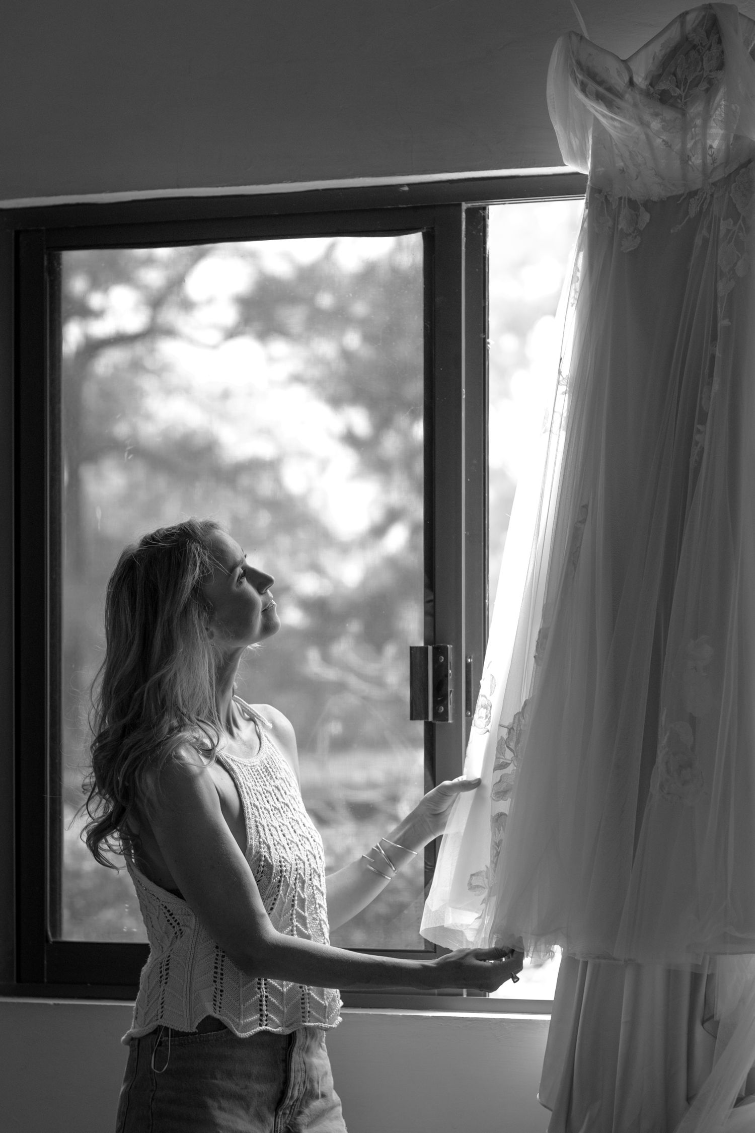 A woman admires a hanging wedding dress by a window in black and white.
