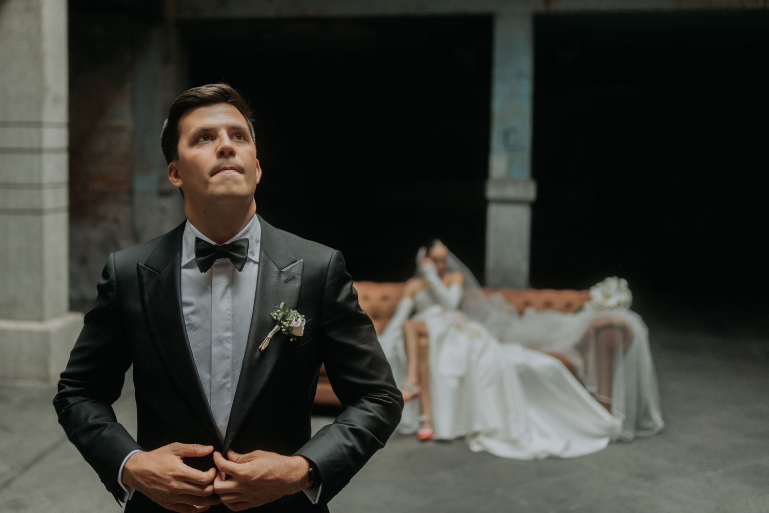 Elegant groom in bow tie and suit poses in foreground with bride seated in background.