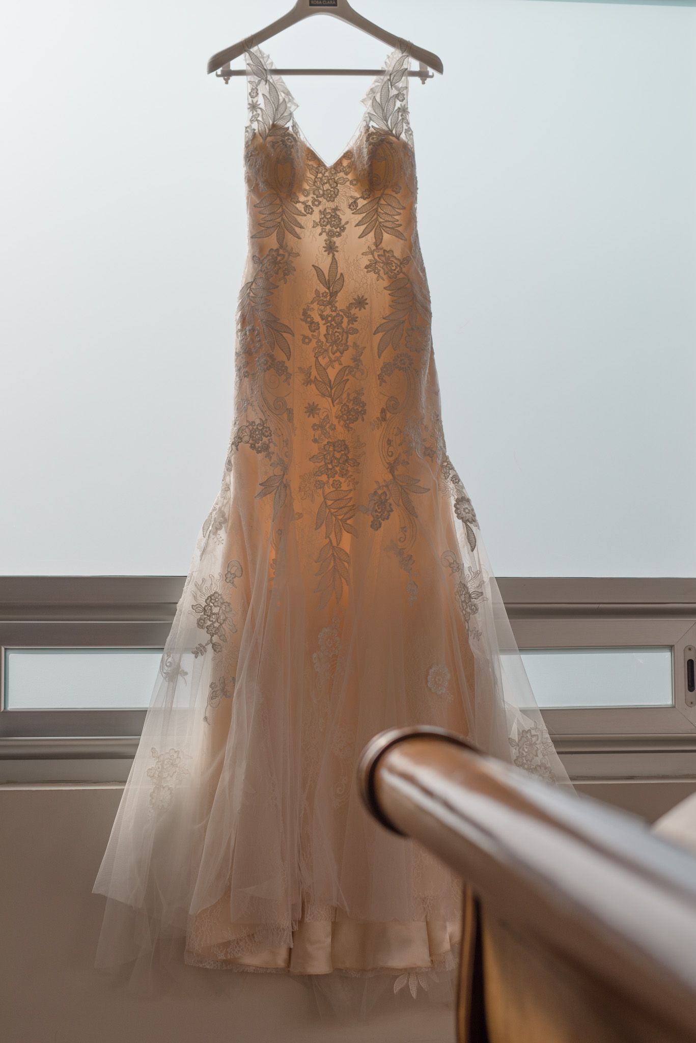 Elegant champagne-colored wedding dress with lace detailing hanging from wooden hanger against white wall.