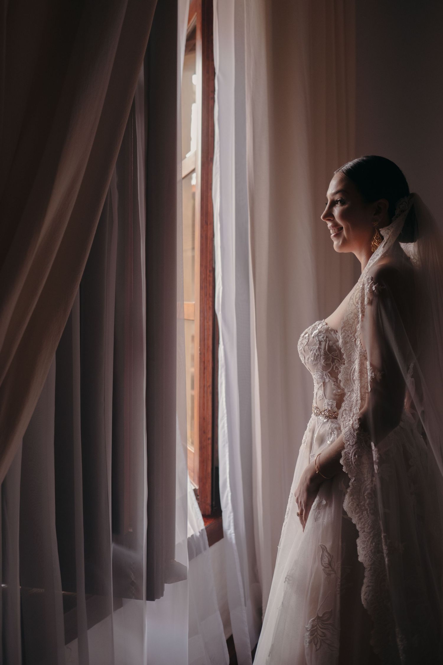 A silhouette by a window shows a wedding dress with intricate lace details and flowing fabric.