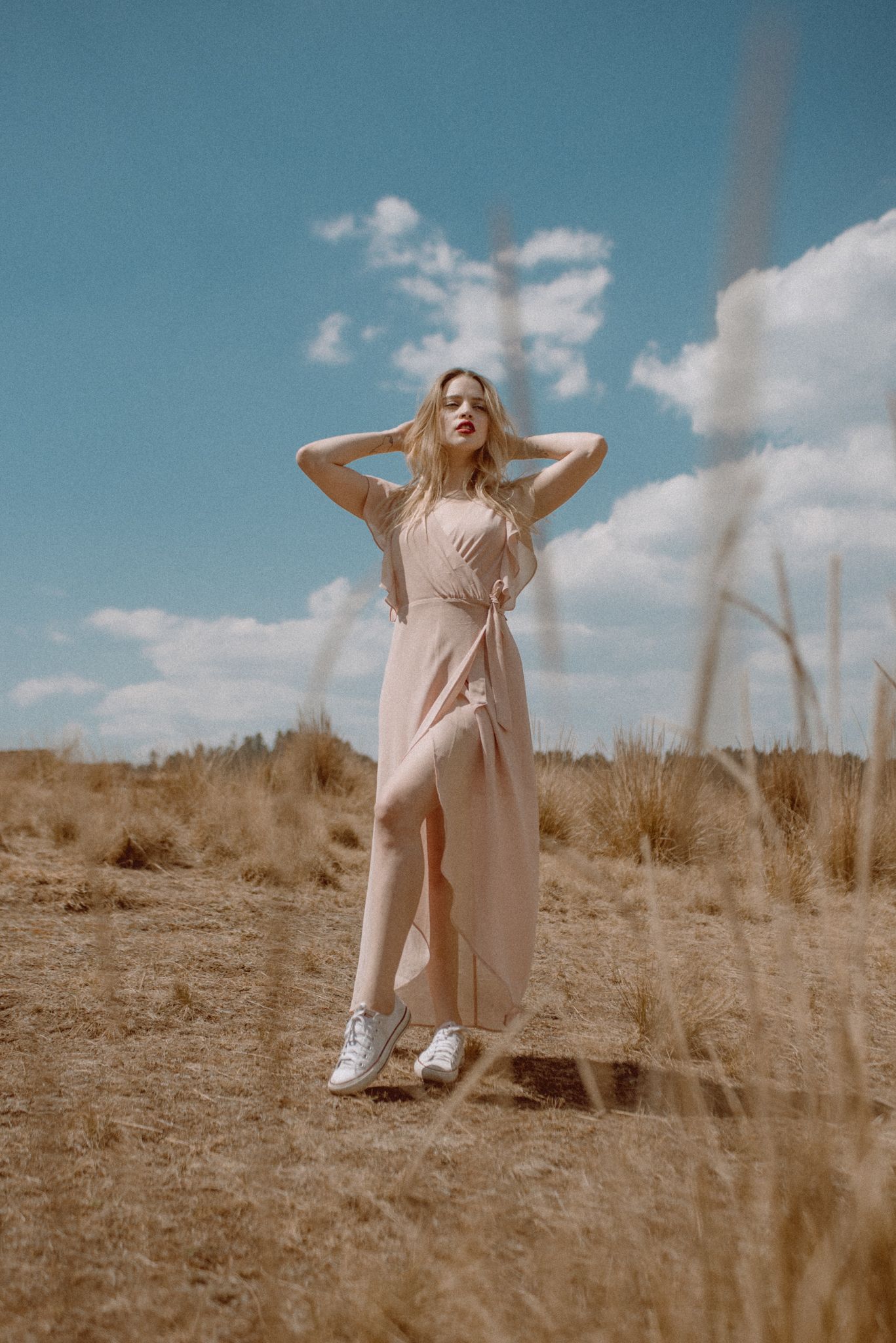 Woman in flowing dress stands among dry grass against bright blue sky.