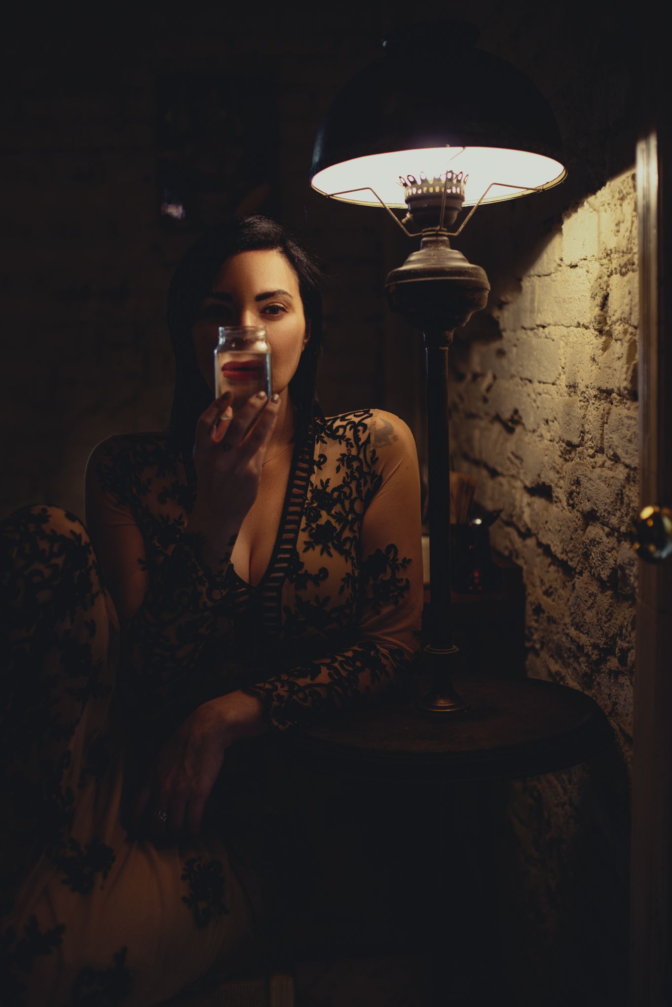 A dark moody portrait of someone in a black lace dress sitting beneath a vintage lamp in a dimly lit room.