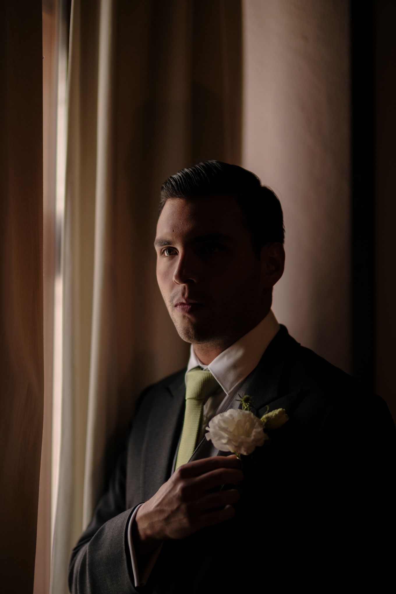 Classic groom portrait by window light wearing dark suit with light green tie and white boutonniere.