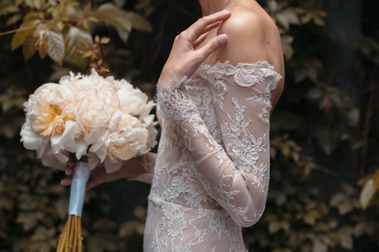 Elegant bride holding a peach-colored peony bouquet while wearing an intricate lace wedding gown.