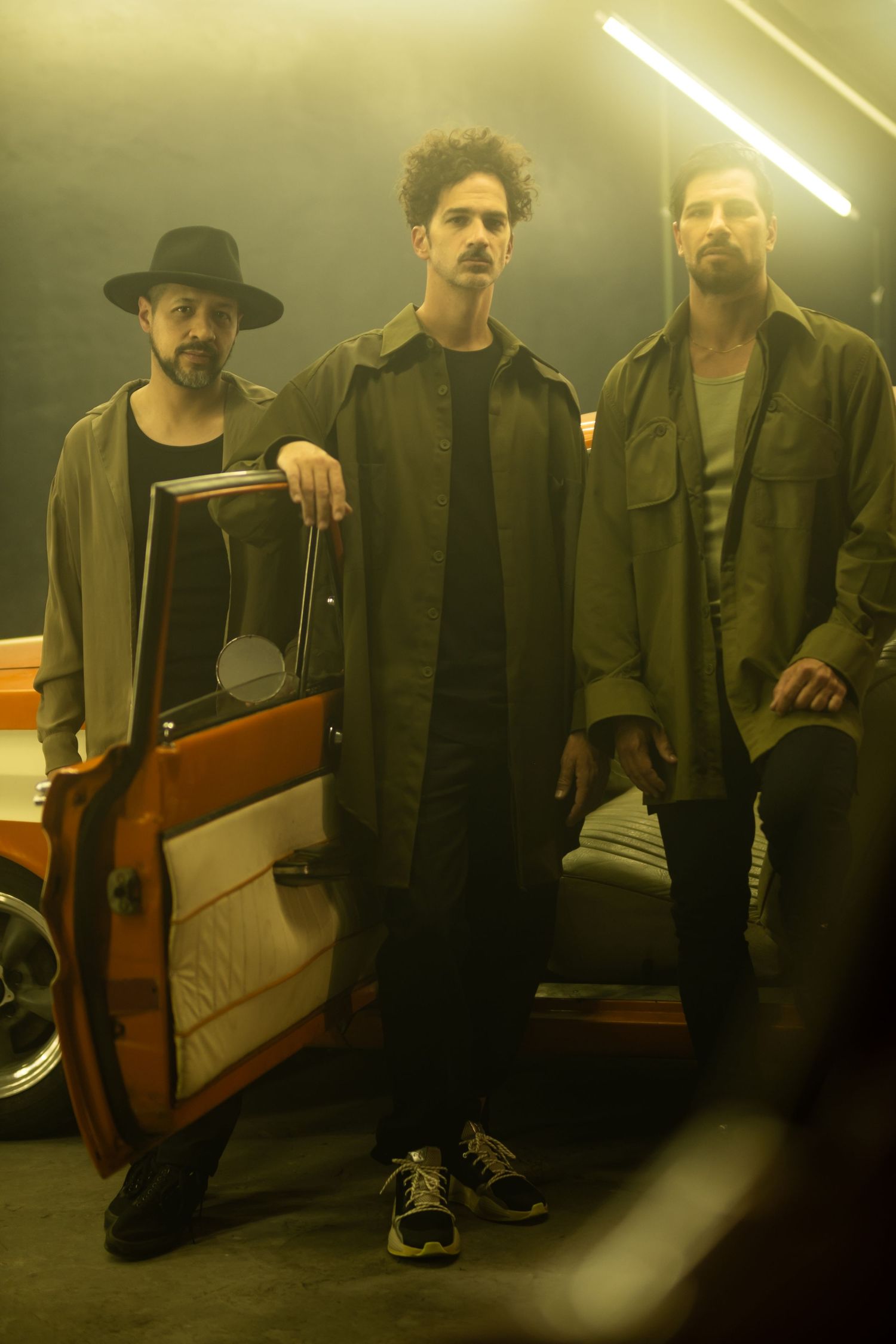 Three musicians pose together in a dimly lit setting with moody yellow lighting and an open car door visible.