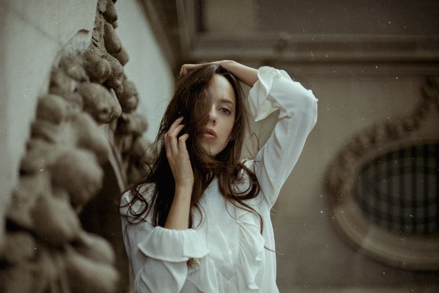 Moody portrait in white blouse against stone architecture in soft lighting.