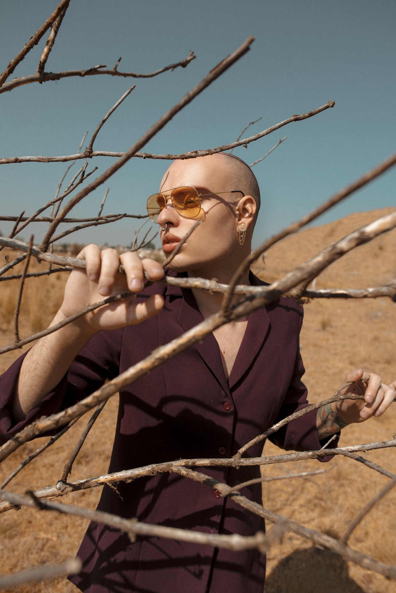 Subject in purple jacket seen through bare branches wearing sunglasses in desert setting.