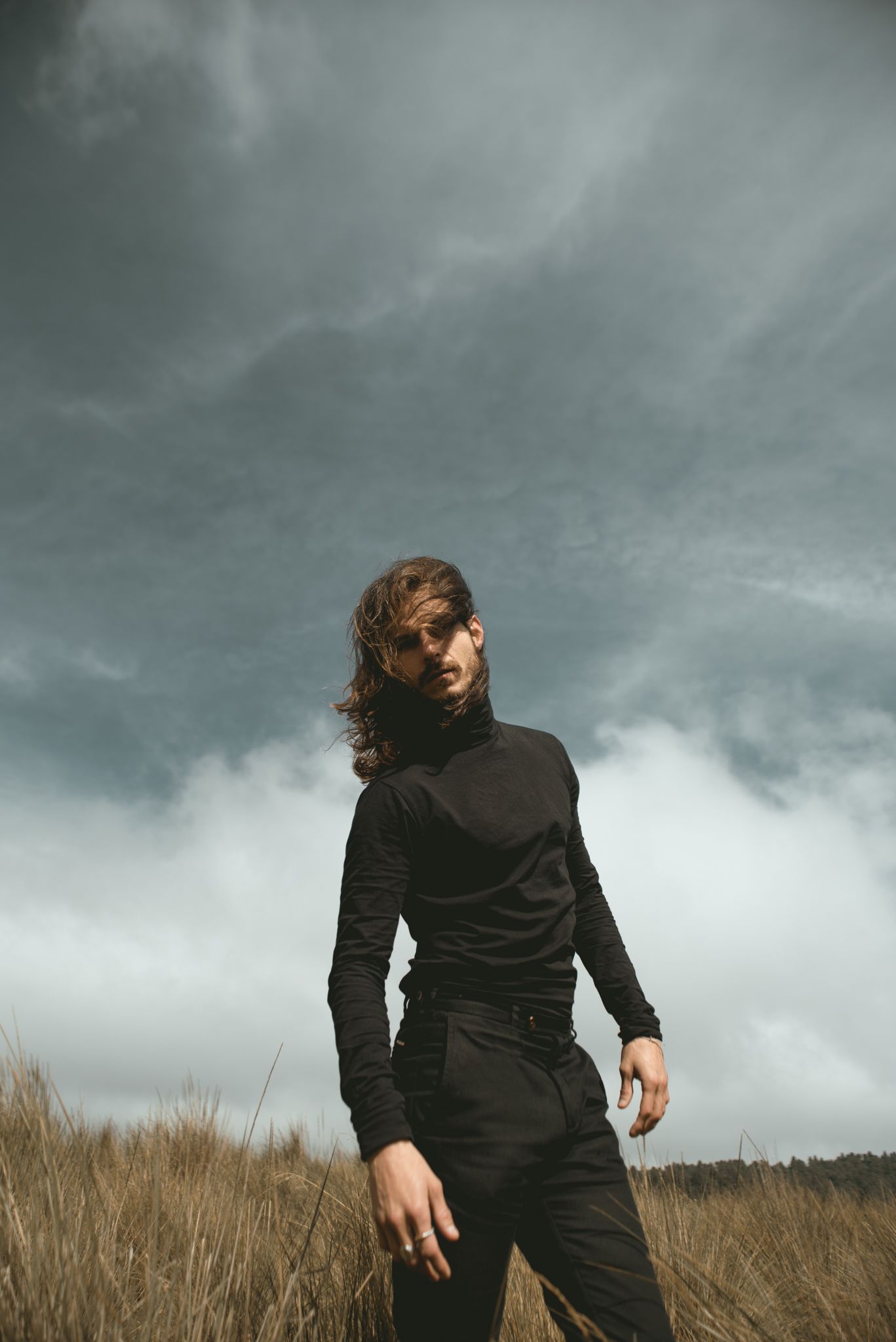 Figure in black outfit standing in field under moody storm clouds.