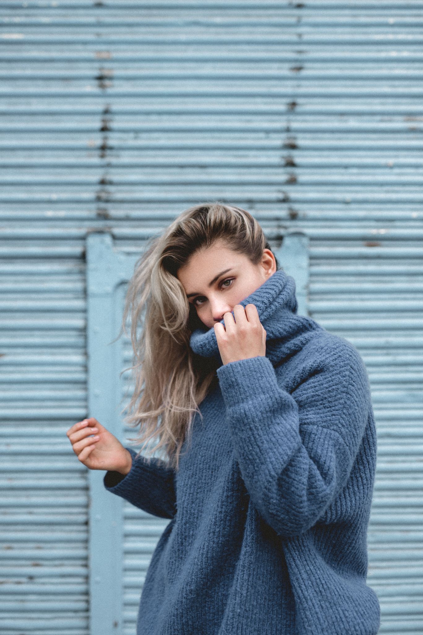 Cozy winter portrait with oversized gray turtleneck sweater against industrial blue background.