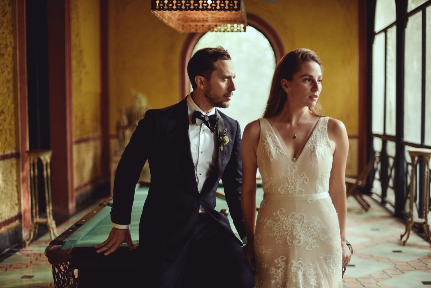 Elegant wedding couple in formal attire pose in vintage corridor with dramatic lighting and ornate architecture.