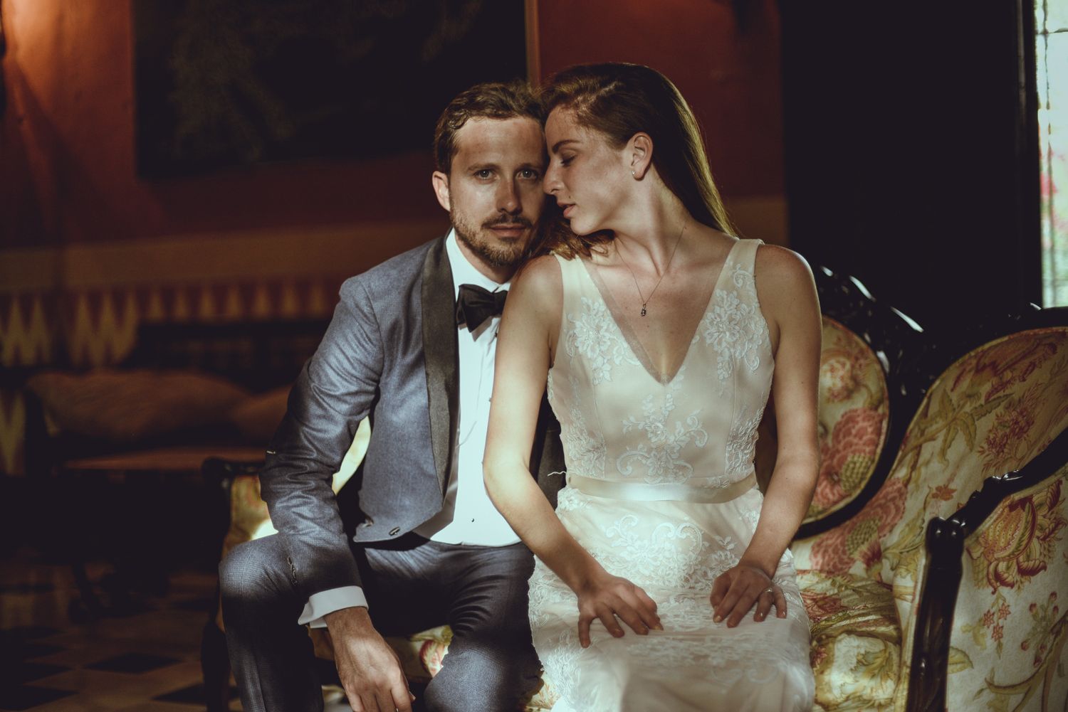 A couple in formal wedding attire shares an intimate moment while seated on an antique sofa in warm lighting.