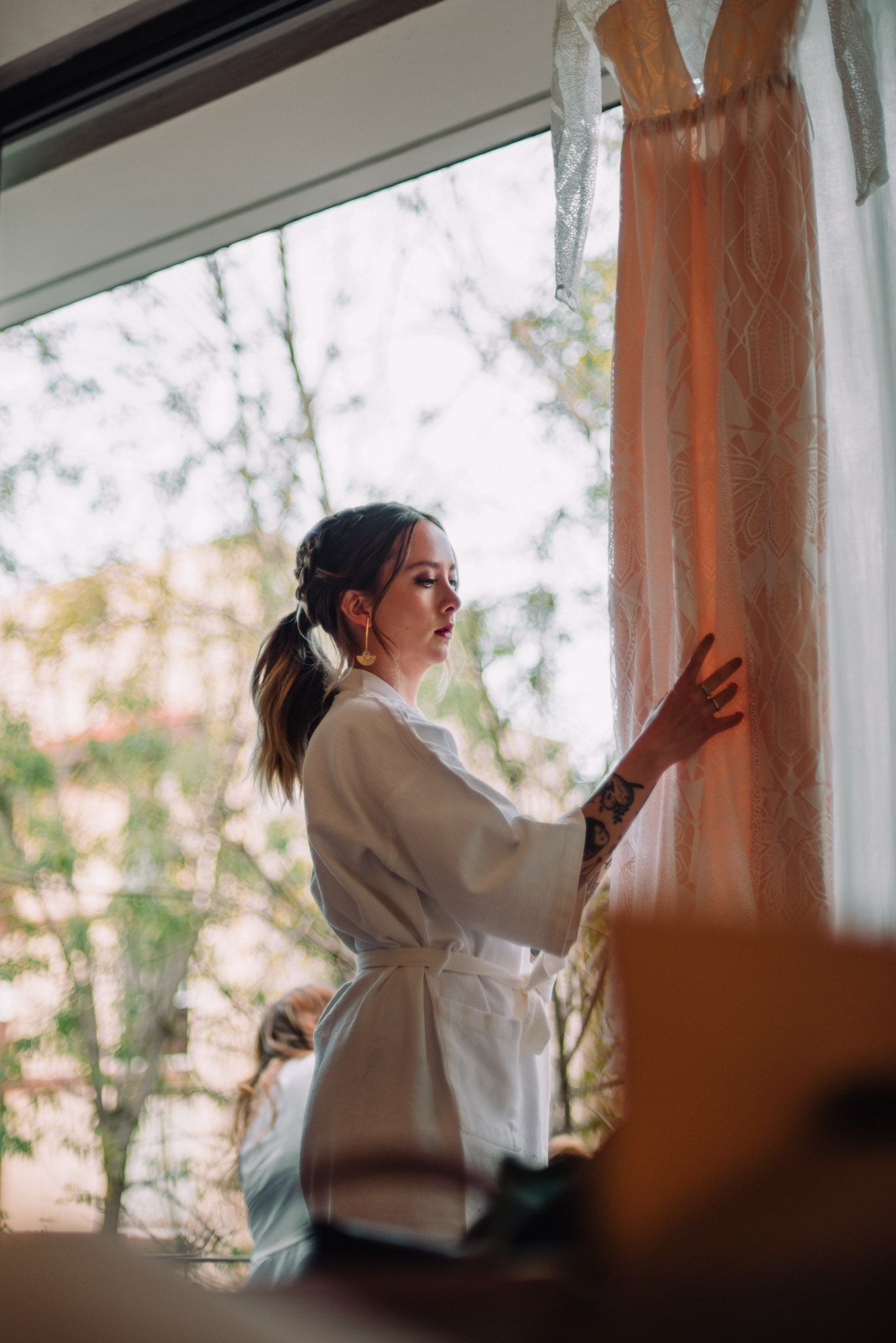 A person in a white silk robe stands by a window preparing for a wedding ceremony in soft morning light.