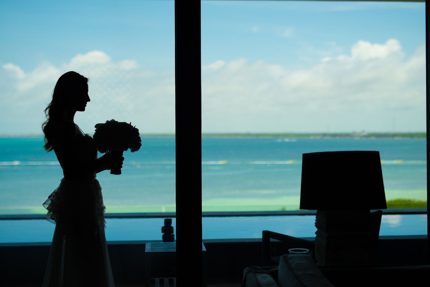 A silhouetted figure holds a bouquet against a bright window overlooking turquoise ocean waters and blue sky.