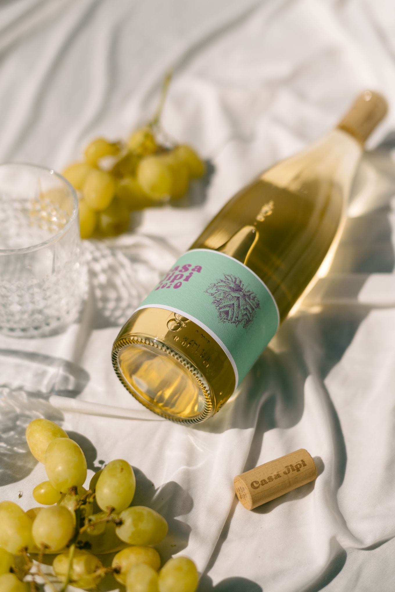 Wine bottle and fresh green grapes lying on white linen cloth in bright sunlight with soft shadows.