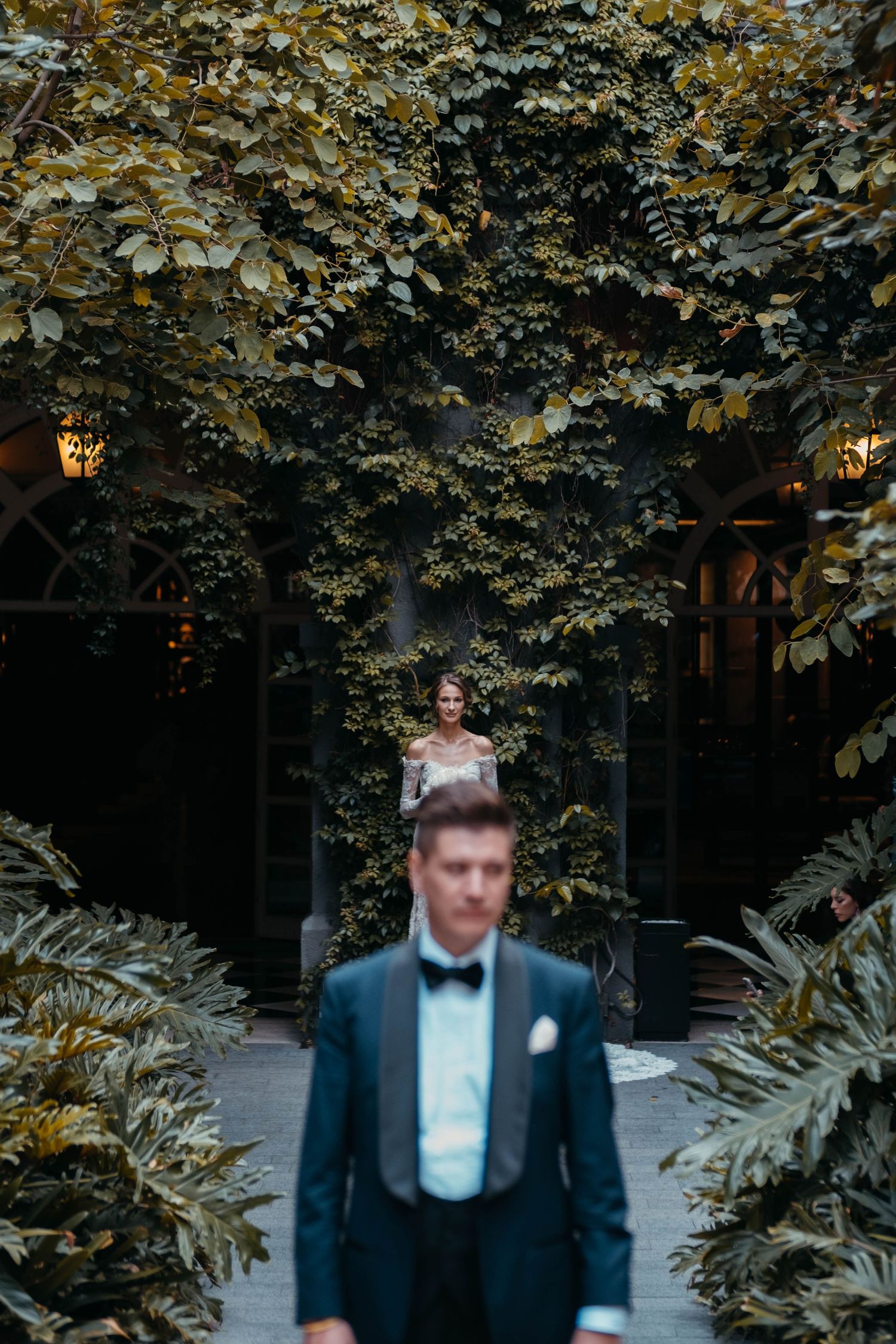 Wedding couple in formal attire posed among lush garden foliage.