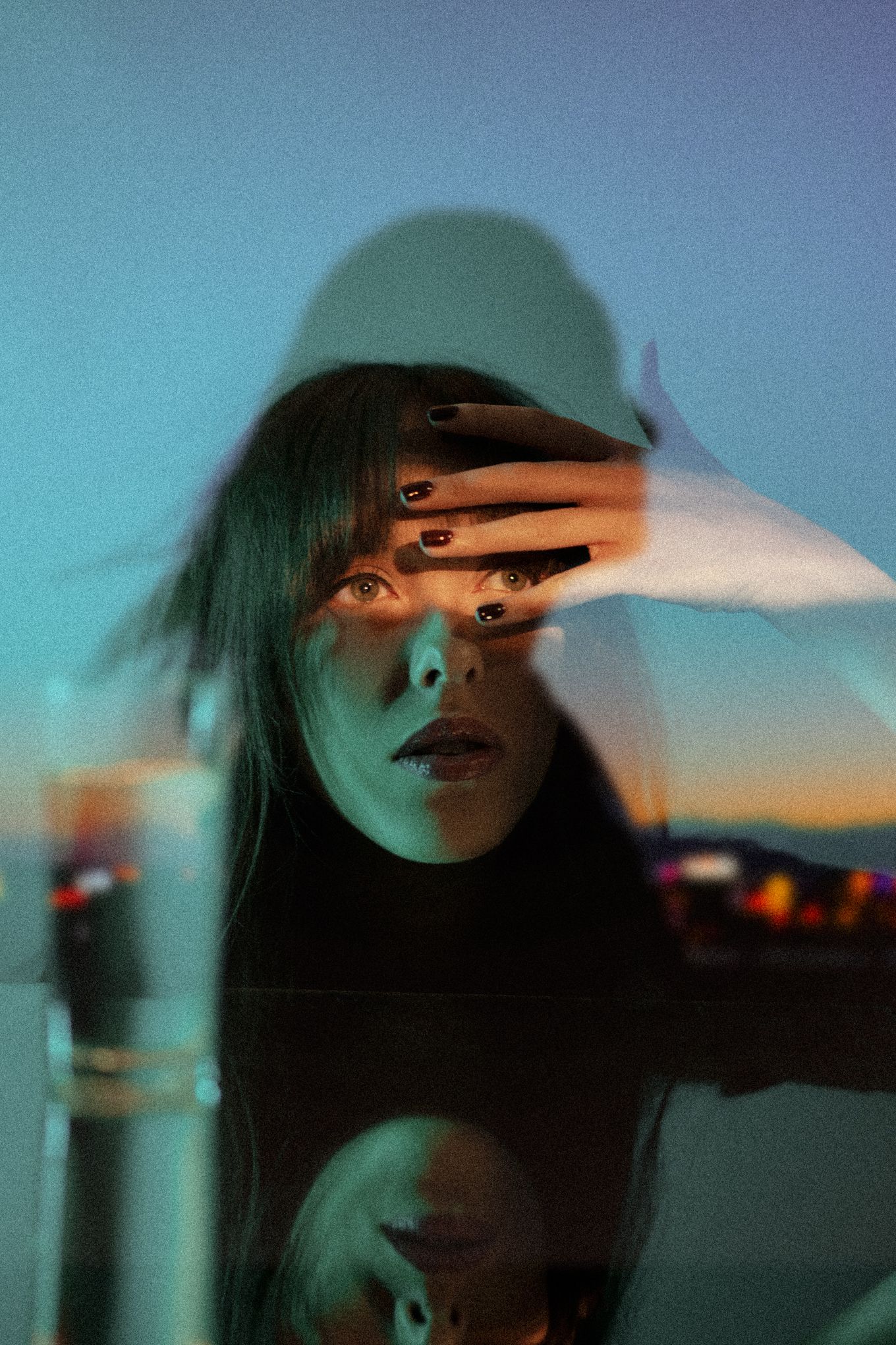 Atmospheric portrait with hand partially covering face against sunset sky reflection.
