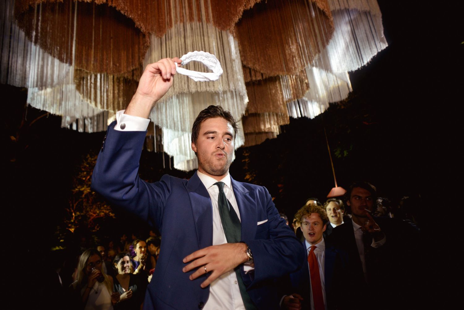 A person in a navy suit holds up a glass for a toast under elegant chandelier lighting.
