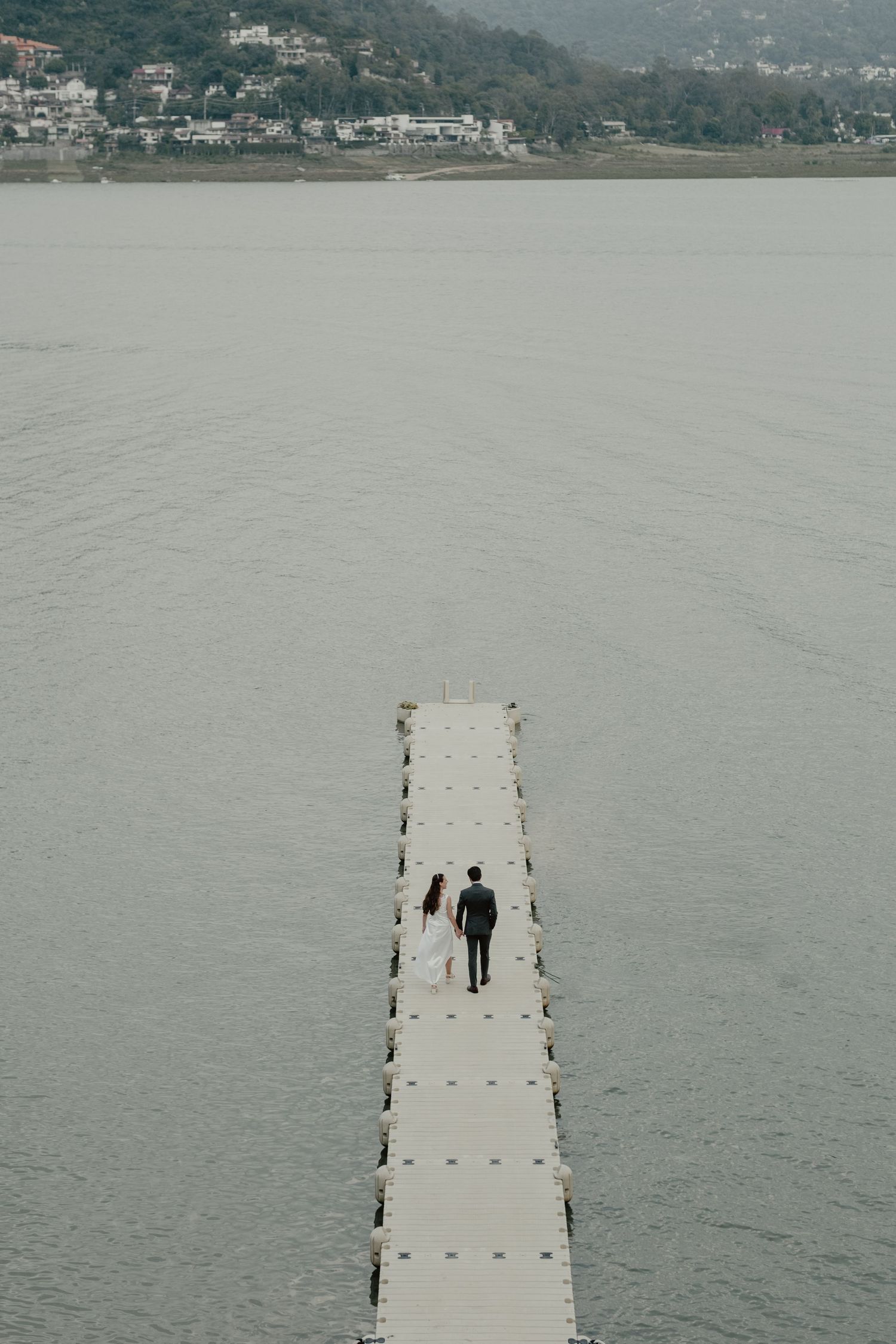Wedding couple walking on long pier over misty lake with mountain backdrop.