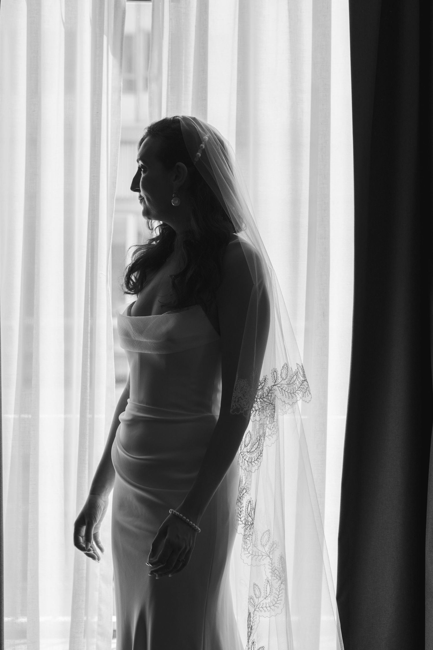 Silhouette of bride in fitted wedding dress standing by sheer white curtained window.