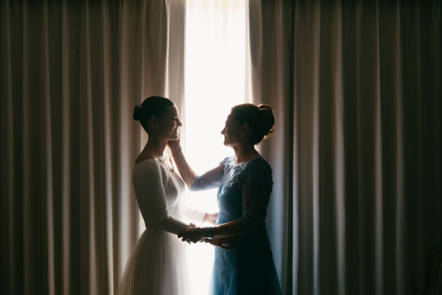 Two silhouettes embrace near a curtained window in a touching wedding day moment.