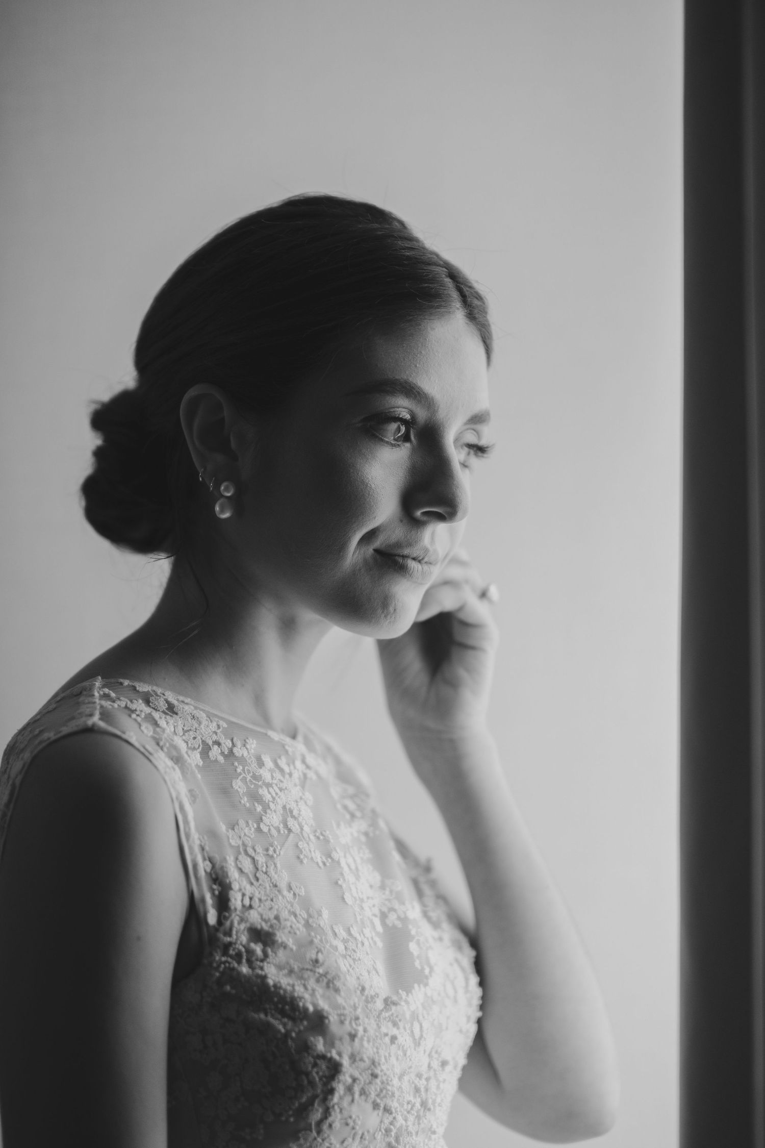 Black and white portrait of bride in lace dress looking thoughtfully by window.