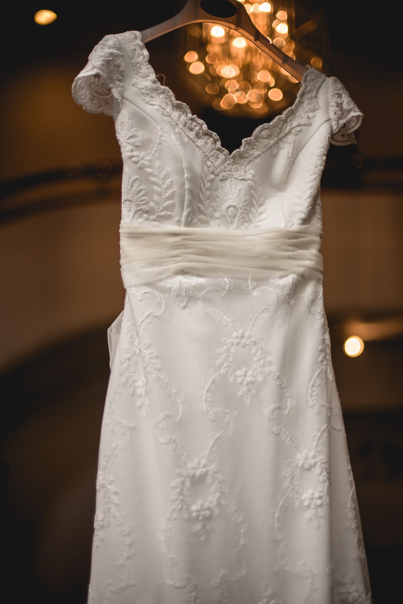 Elegant white wedding dress with lace detail and capped sleeves hanging on display with bokeh lights in background.