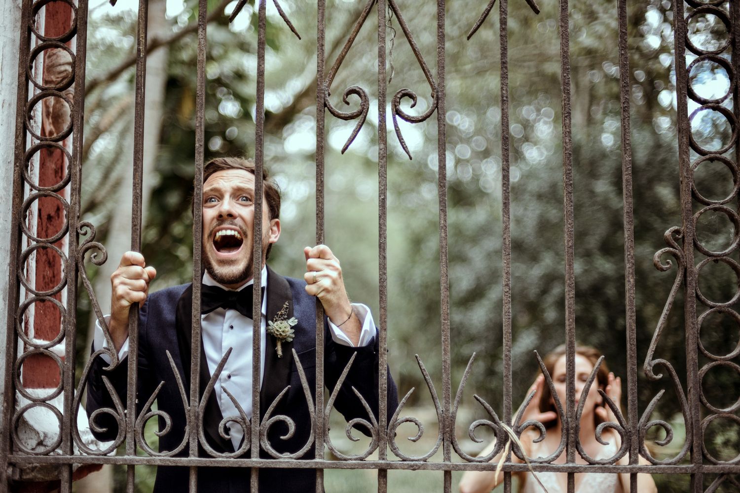 A person in formal attire grasps wrought iron gates while showing playful excitement behind decorative metal bars.