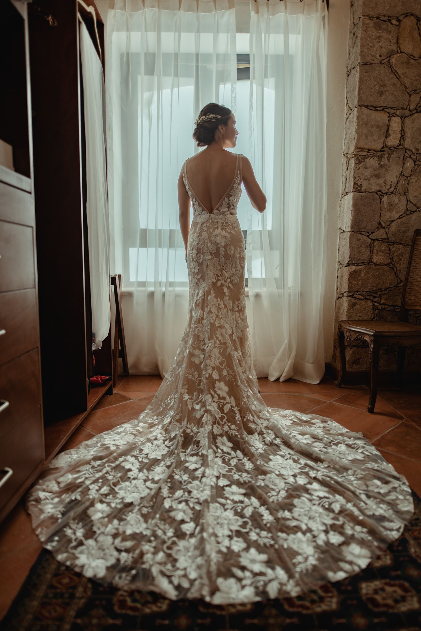 Elegant lace wedding dress with dramatic cathedral train shown in silhouette against sunlit window in rustic stone room.