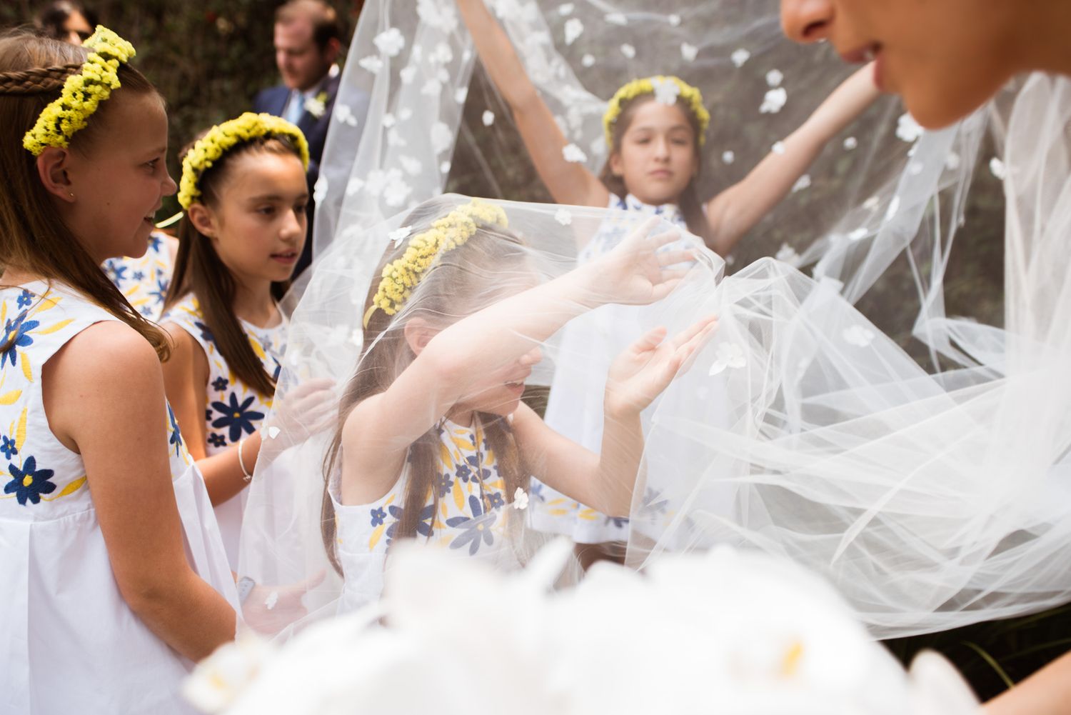 Children in white dresses with yellow headbands celebrate around flowing tulle fabric during a special occasion.