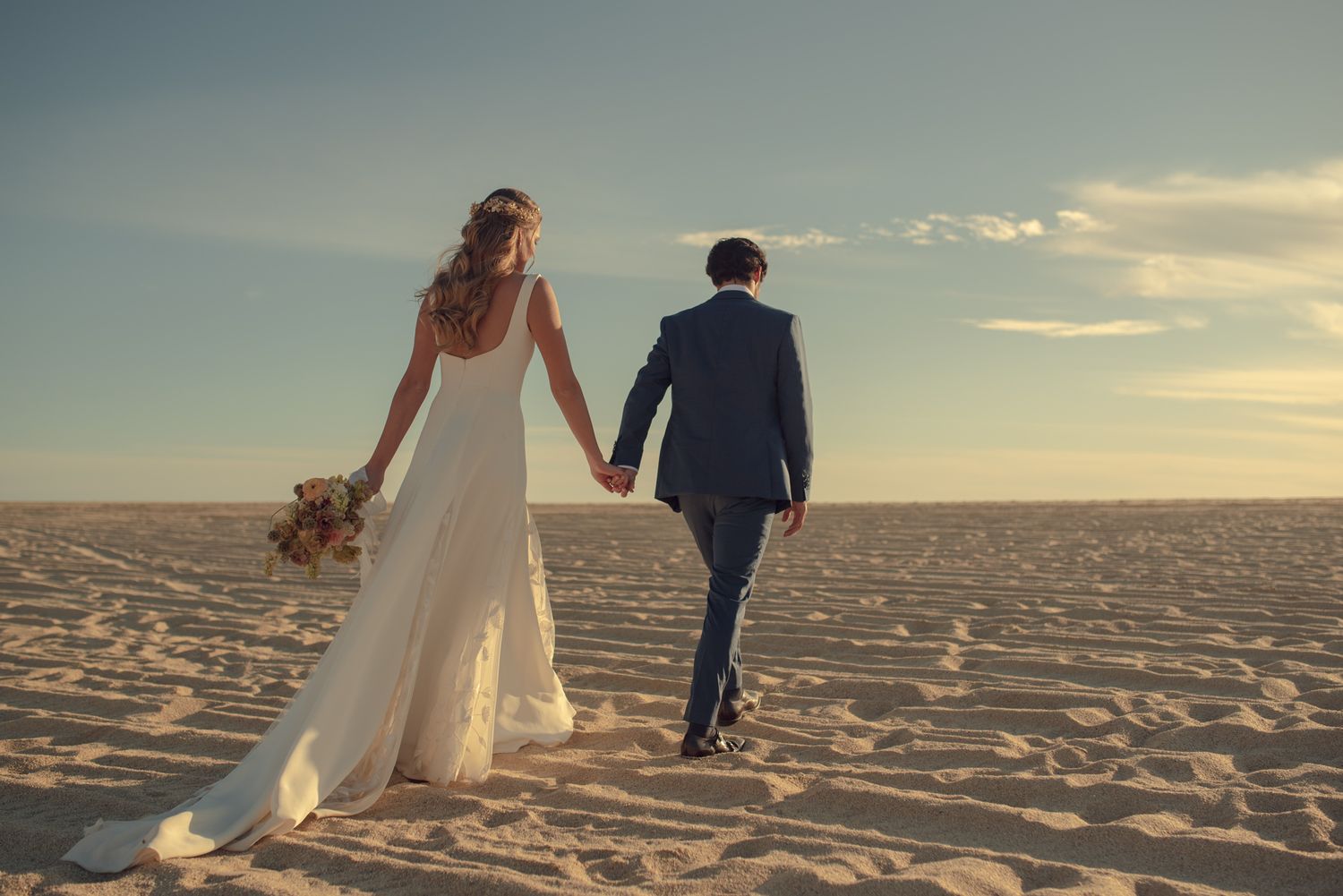 Wedding couple walking hand in hand across sandy beach at sunset holding bouquet with flowing white dress.