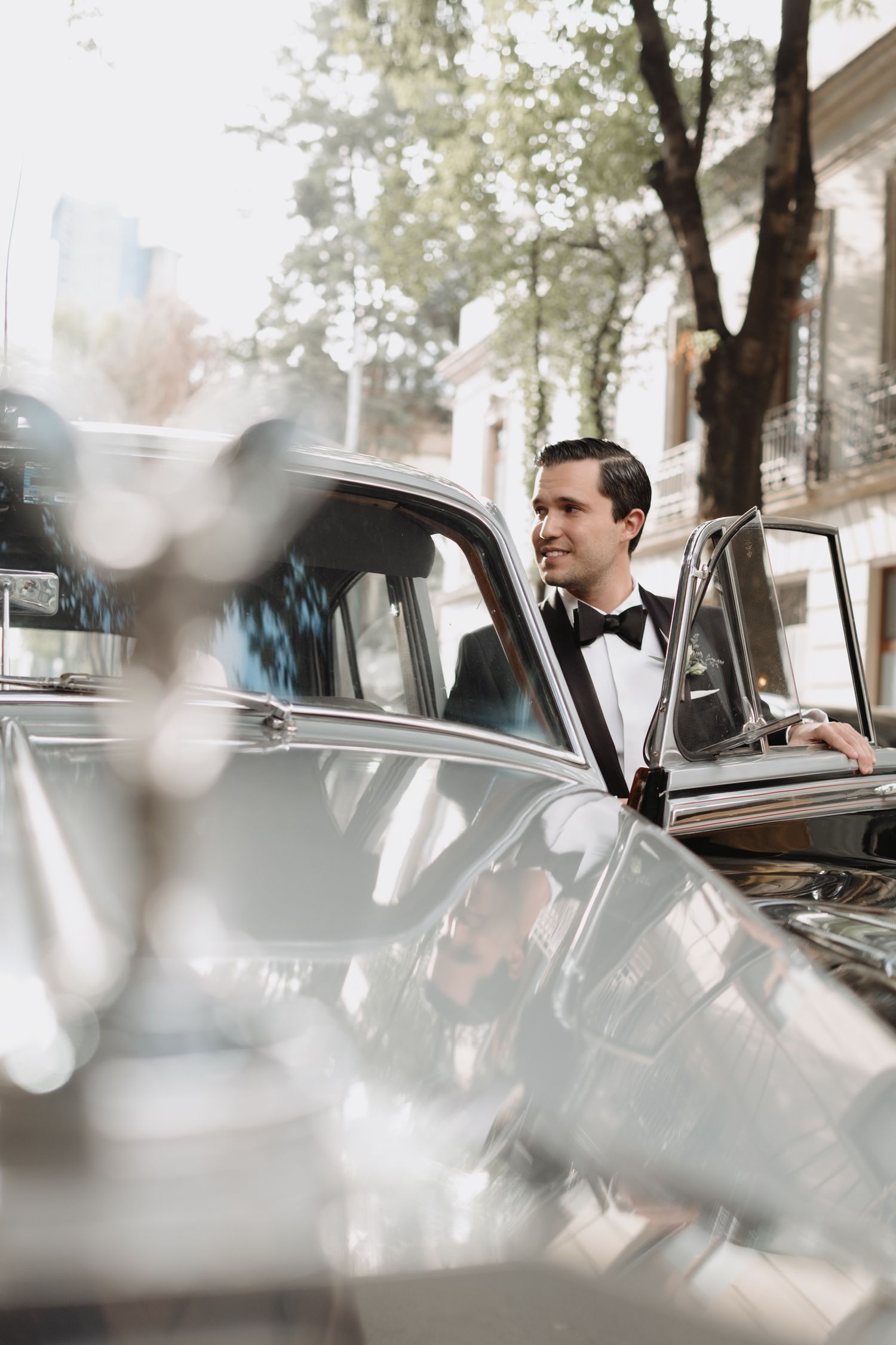 A person in formal attire leans out of a classic vintage car on a tree-lined street.