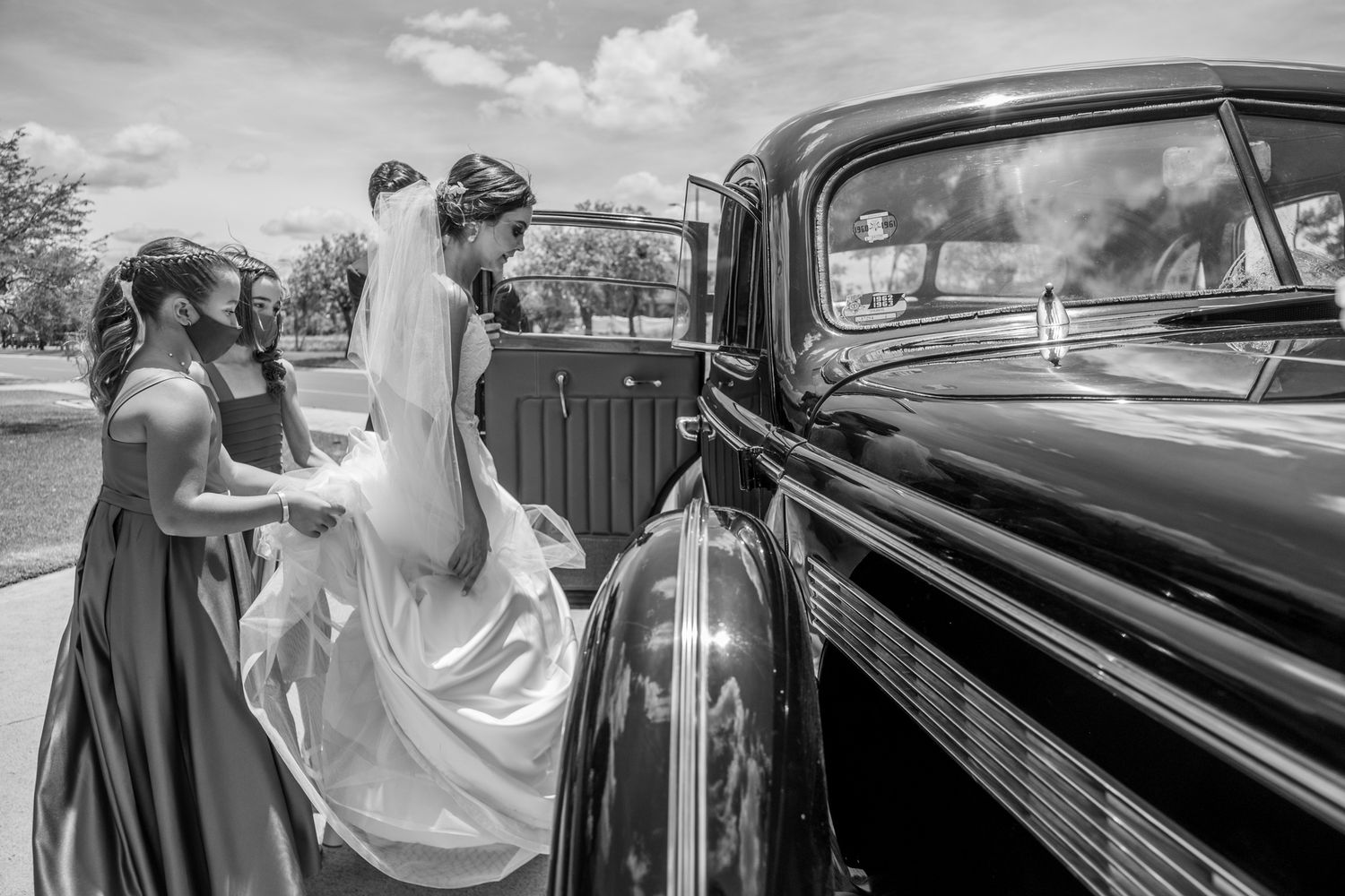 Vintage black car at a wedding ceremony with flowing white dress and bridesmaid preparing for transport.