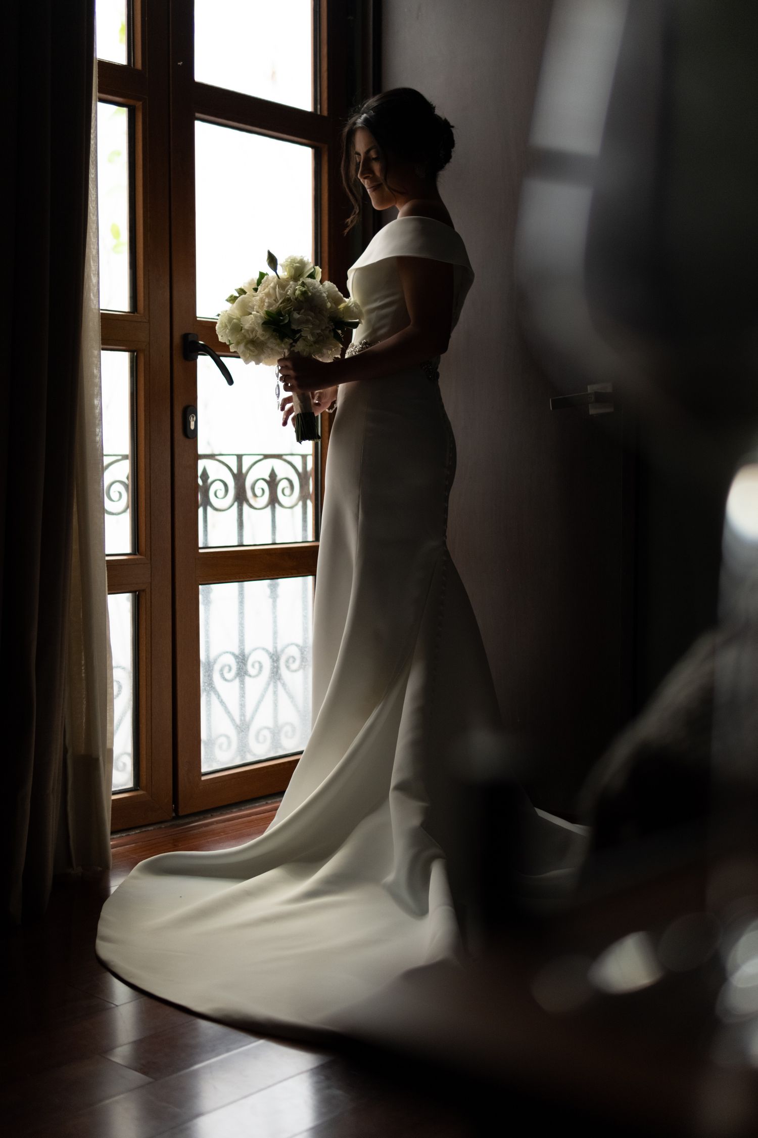 Silhouette of a bride in an elegant white wedding dress holding a bouquet while standing by a window with ornate railings.