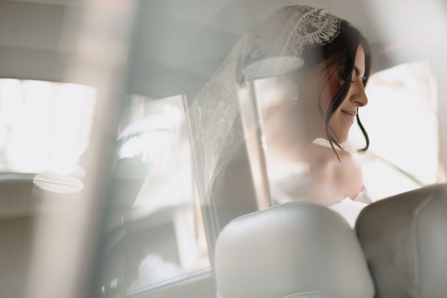 Ethereal bridal portrait through car window with soft natural light and flowing veil.