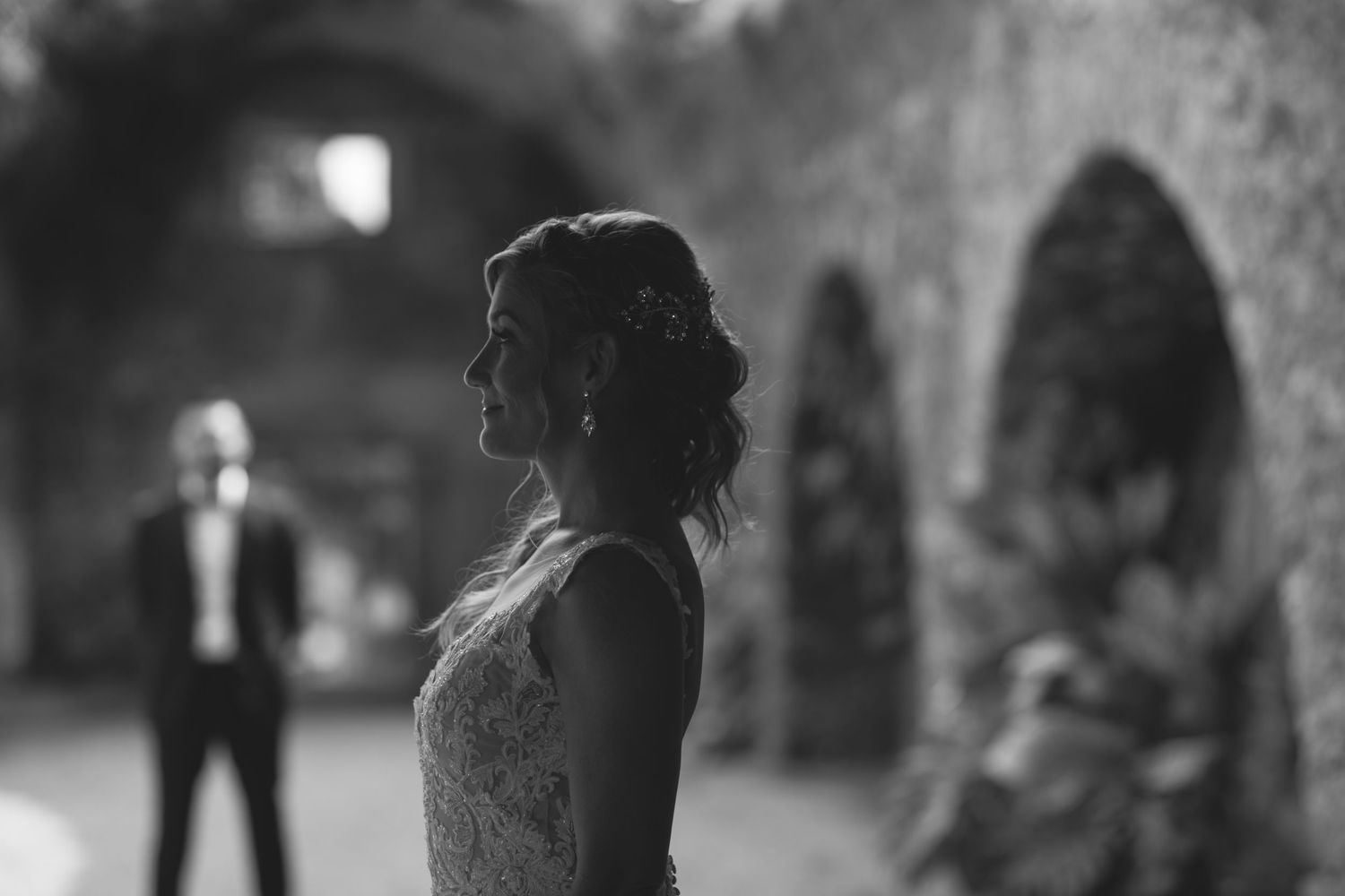A black and white silhouette of a bride in lace dress under stone arches, with a distant figure in background.