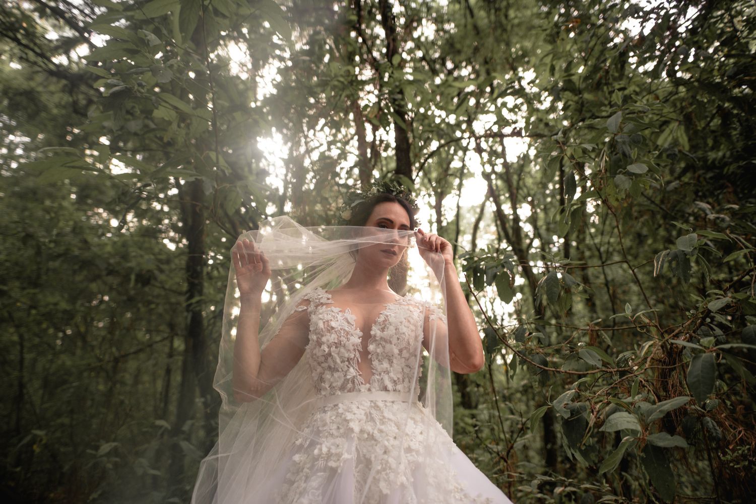 A person in a detailed white lace wedding dress poses in a misty forest setting with sunlight filtering through trees.