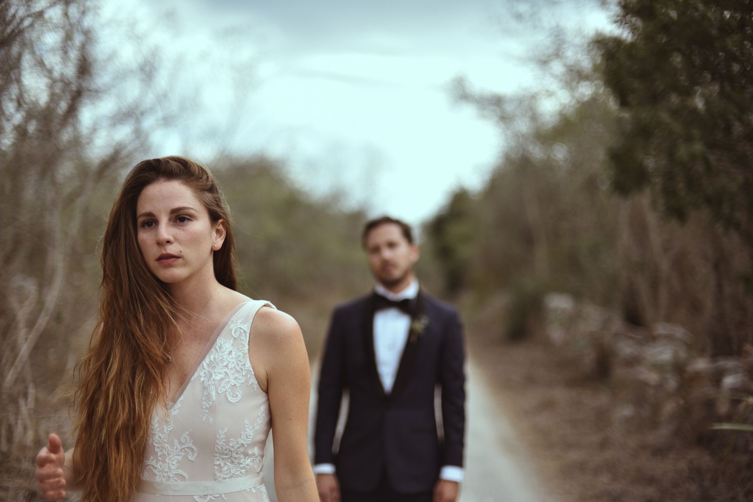 A newlywed couple poses on a path with focus on the bride in a white dress while the groom stands in background.