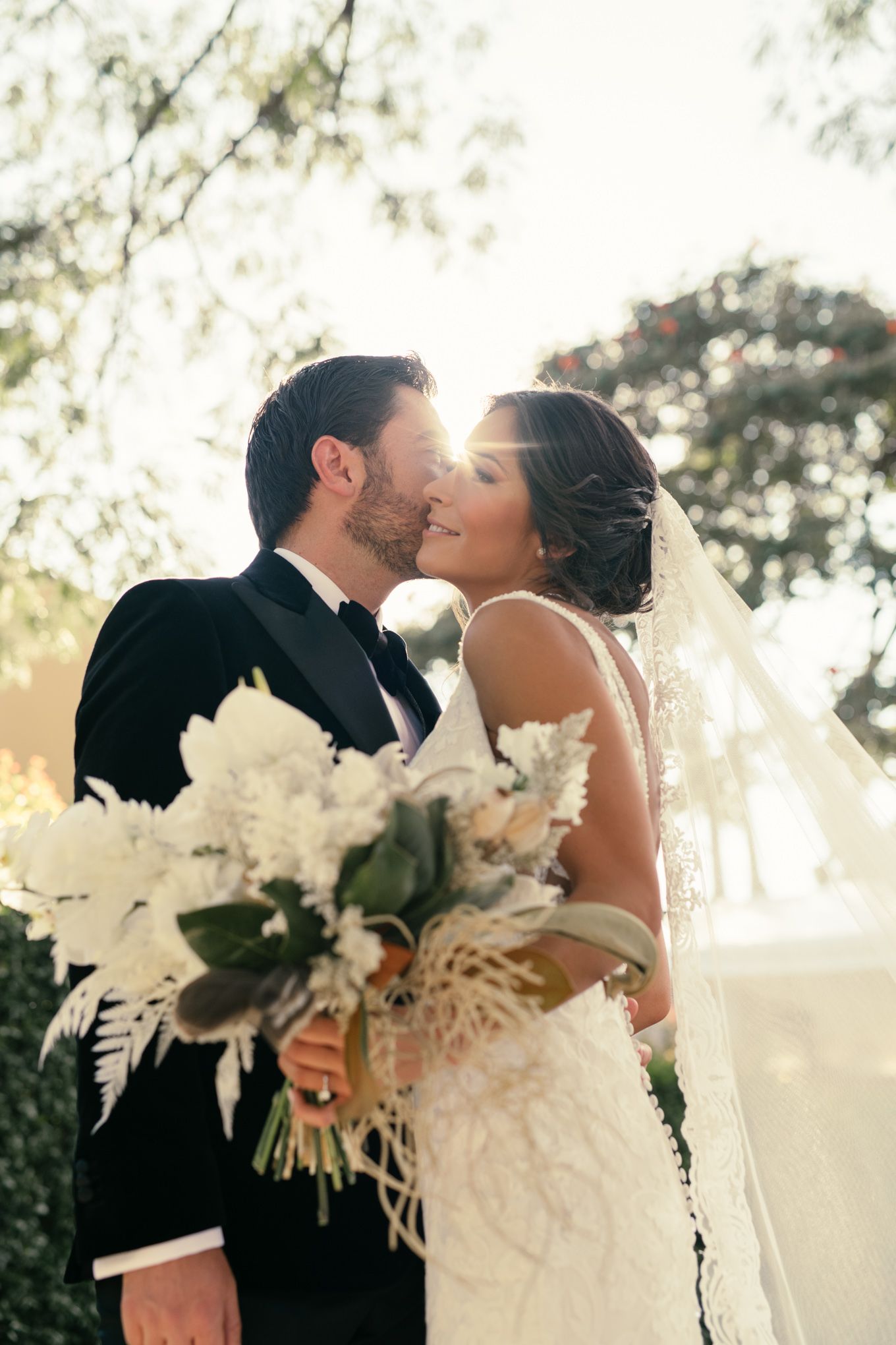 Romantic wedding kiss captured in sunlit outdoor setting with white floral bouquet.