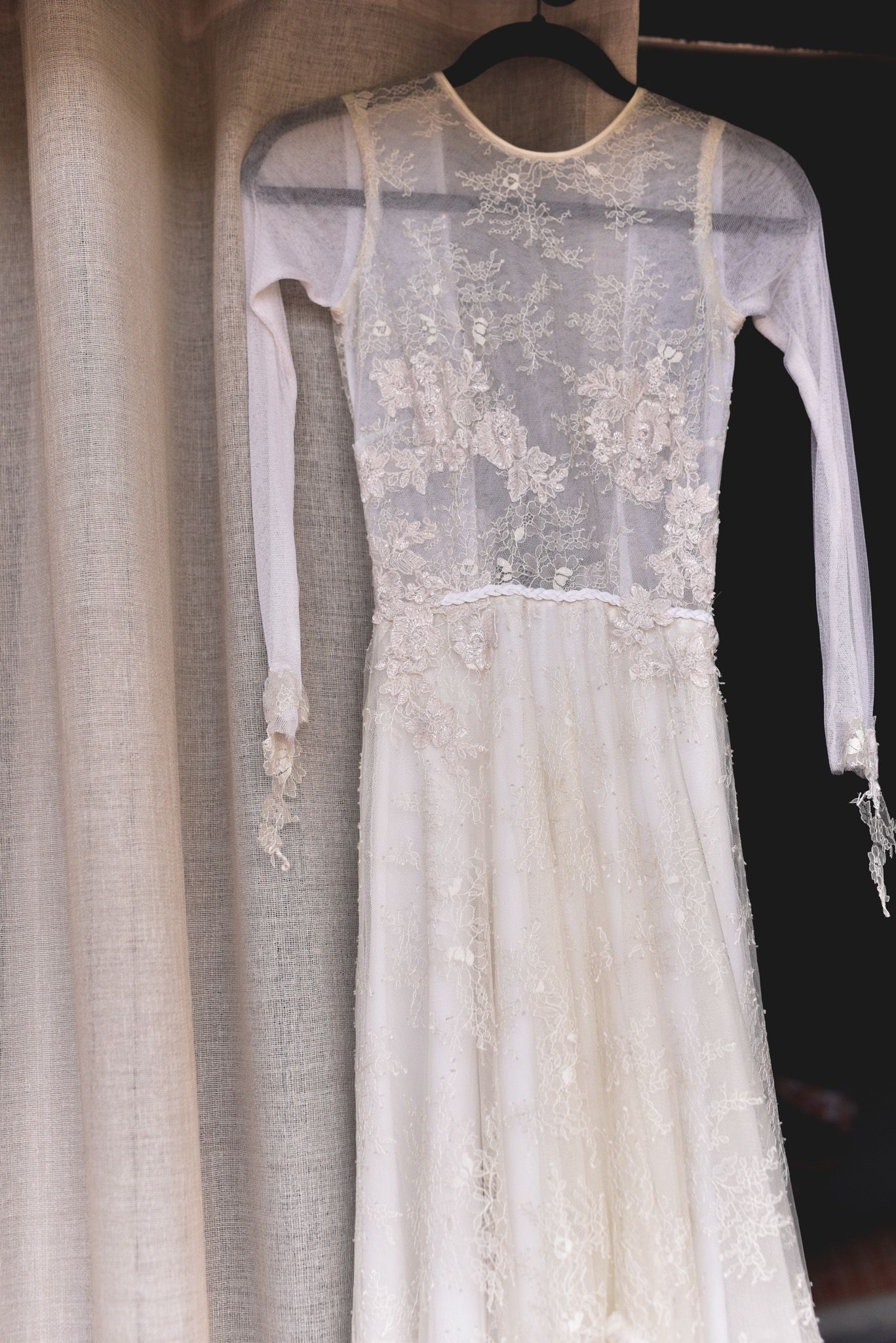 A delicate lace wedding dress with long sleeves hanging against a light background.