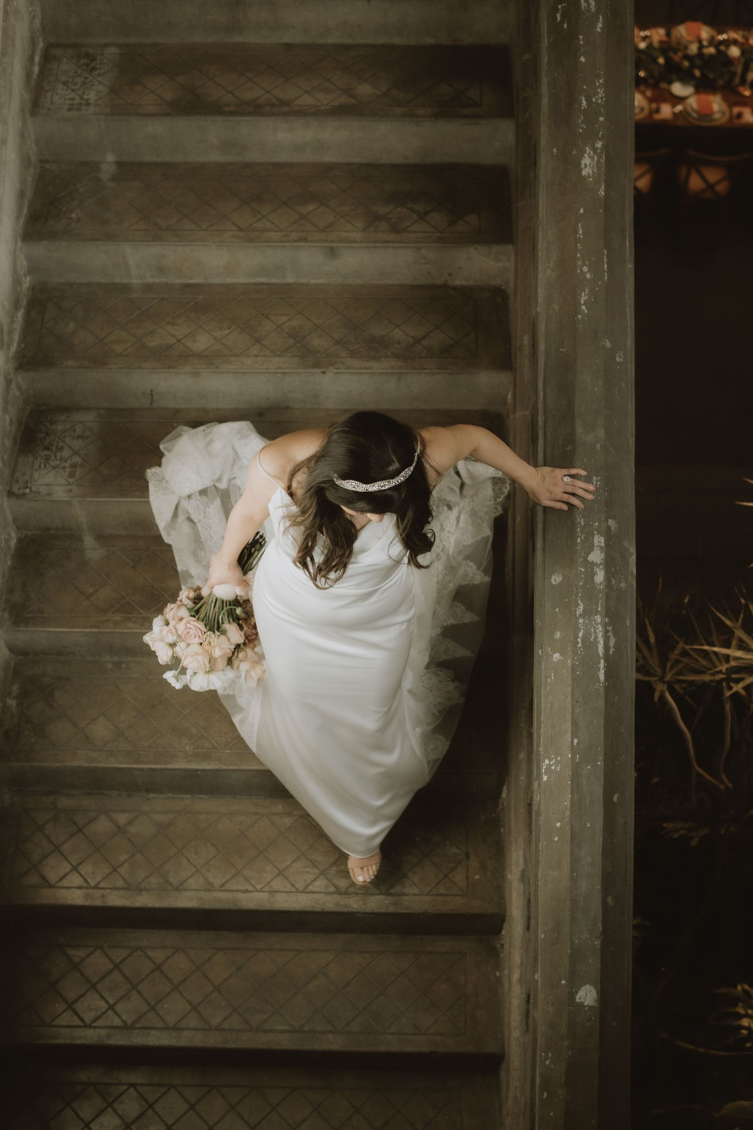 Overhead view of bride in flowing white dress descending vintage stairs while holding bouquet.